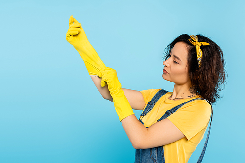 brunette woman putting on yellow rubber gloves isolated on blue