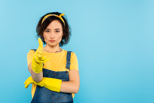 woman in denim overalls and rubber gloves showing gun gesture isolated on blue