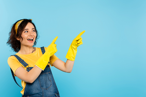 excited woman in yellow rubber gloves looking away and pointing with fingers isolated on blue