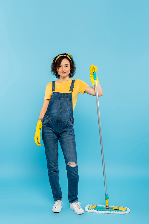 full length view of woman in denim overalls and rubber gloves posing with mop on blue