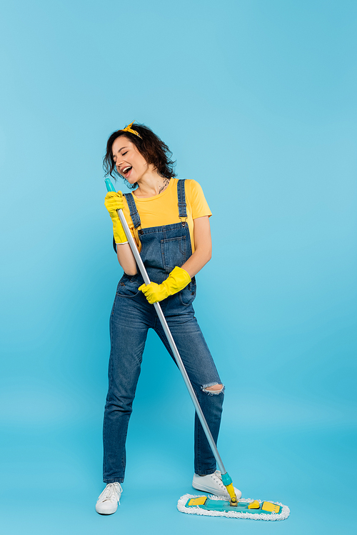 cheerful housewife in denim overalls and rubber gloves singing with mop on blue