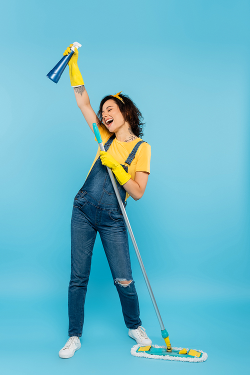 excited woman in rubber gloves and denim overalls singing with detergent and mop on blue