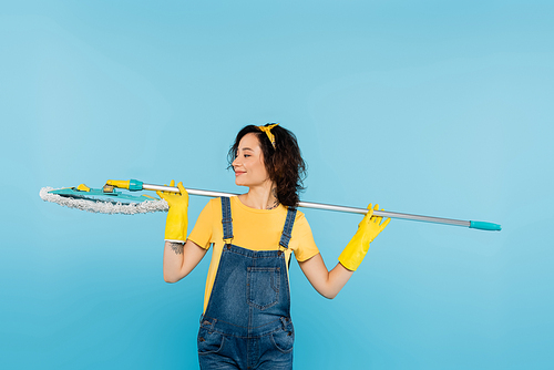 smiling woman in rubber gloves and denim overalls standing with mop isolated on blue