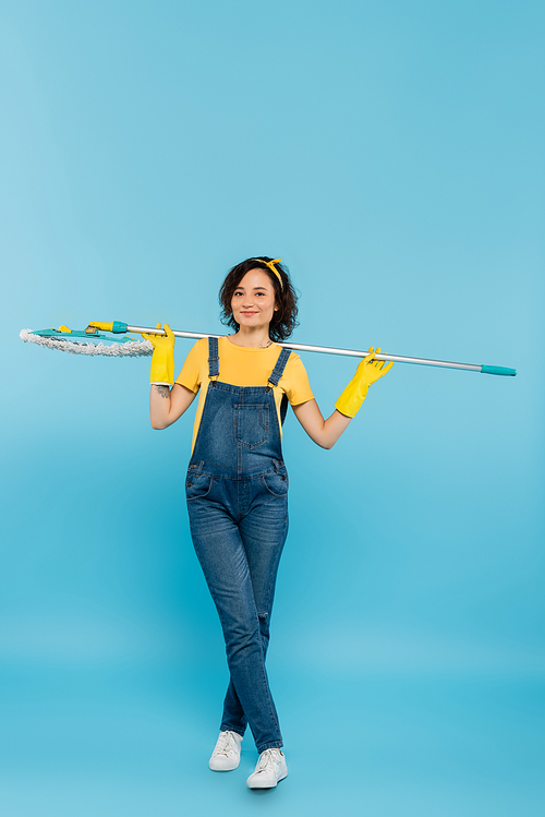 happy housewife in yellow rubber gloves and denim overalls posing with mop on blue