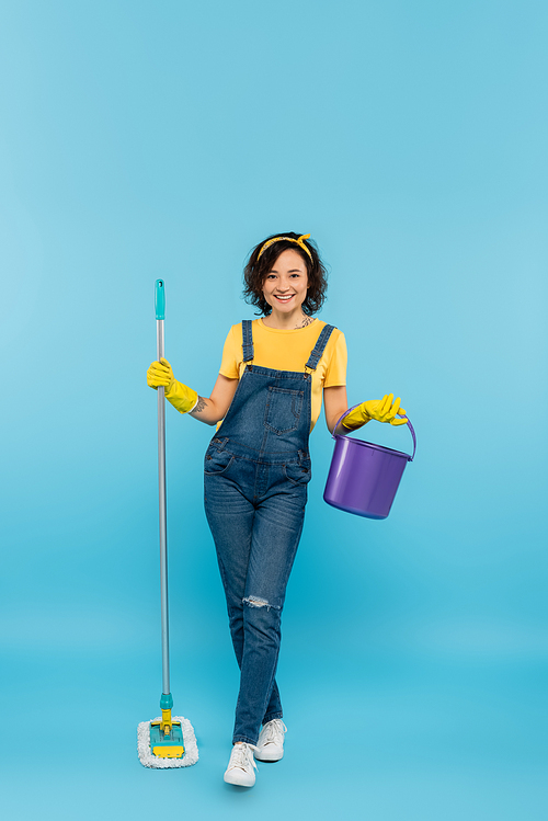 full length view of woman in yellow rubber gloves posing with bucket and mop on blue
