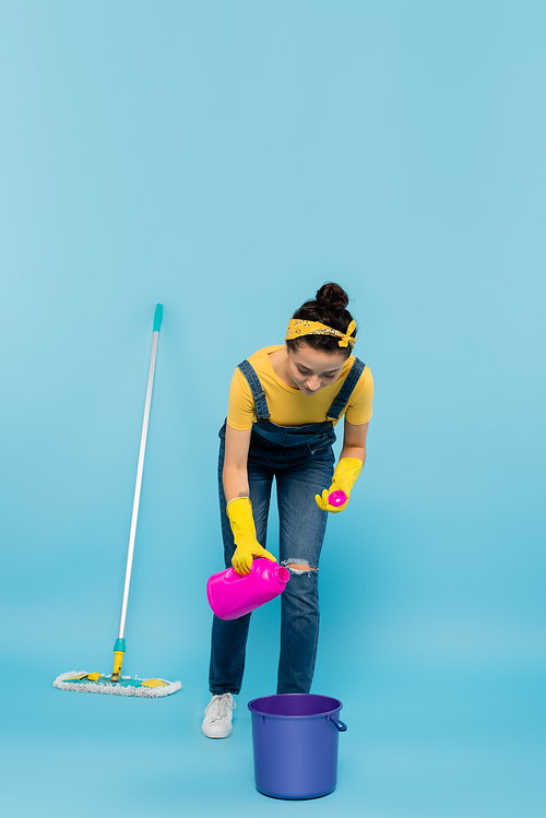 housewife in denim overalls and rubber gloves pouring detergent into bucket near mop on blue