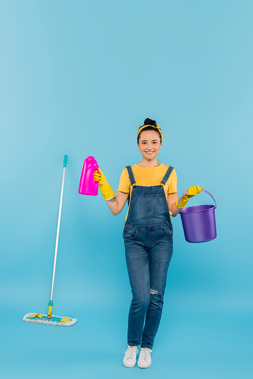 smiling housewife in denim overalls holding detergent and bucket near mop on blue