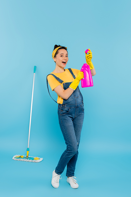 excited woman in denim overalls holding detergent while standing near mop on blue