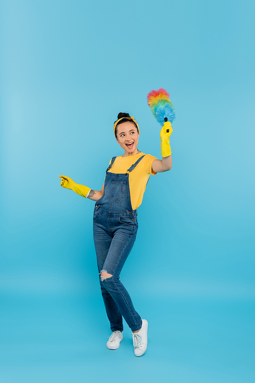 excited housewife in denim overalls waving dust brush while looking away on blue