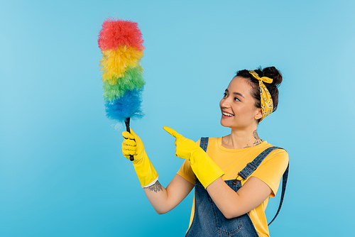 smiling housewife in yellow rubber gloves pointing at colorful dust brush isolated on blue