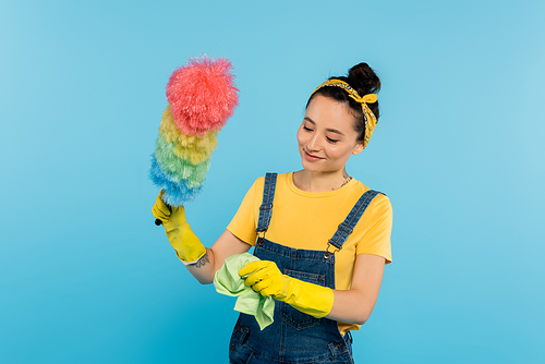 smiling woman in denim overalls holding rag and colorful dust brush isolated on blue