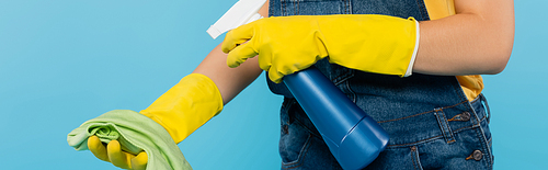 cropped view of woman in rubber gloves spraying detergent on rag isolated on blue, banner