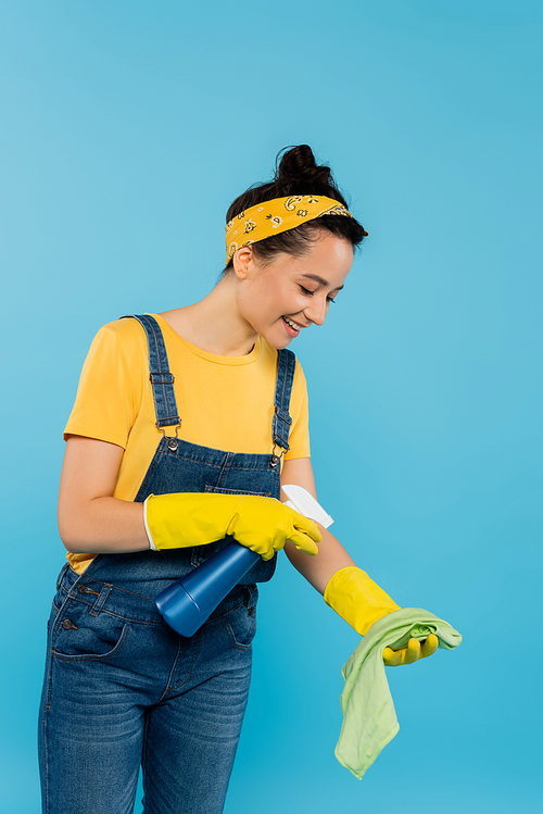 smiling housewife spraying detergent on rag isolated on blue