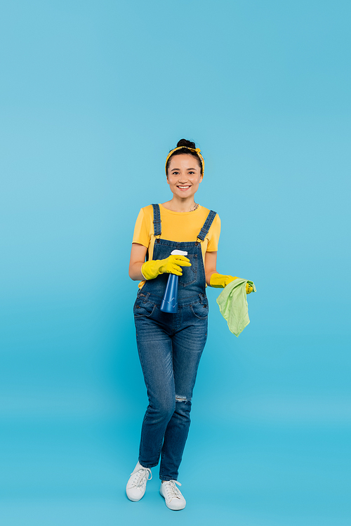 full length view of housewife in denim overalls and latex gloves holding rag and detergent on blue