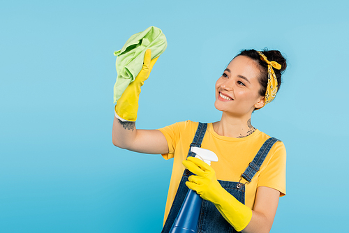 happy woman in yellow rubber gloves holding rag and spray bottle with cleanser isolated on blue