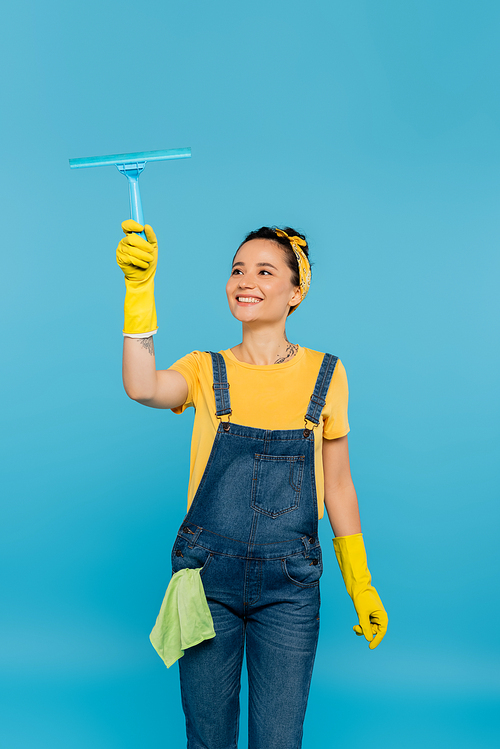 happy housewife with rag in pocket of denim overalls holding window cleaner isolated on blue