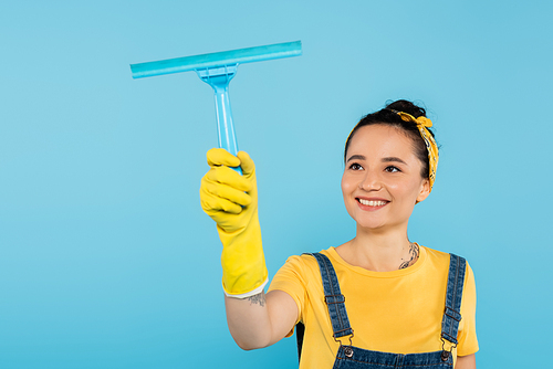 pleased woman in yellow rubber glove holding window cleaner isolated on blue
