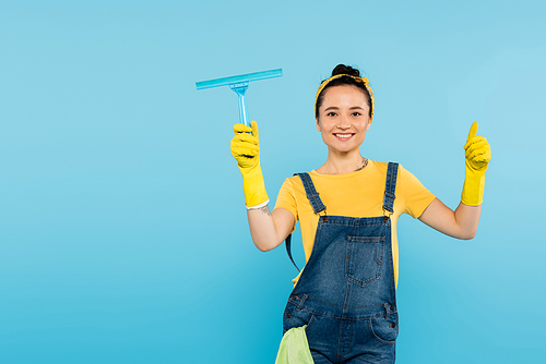 joyful housewife with window cleaner showing thumb up while  isolated on blue