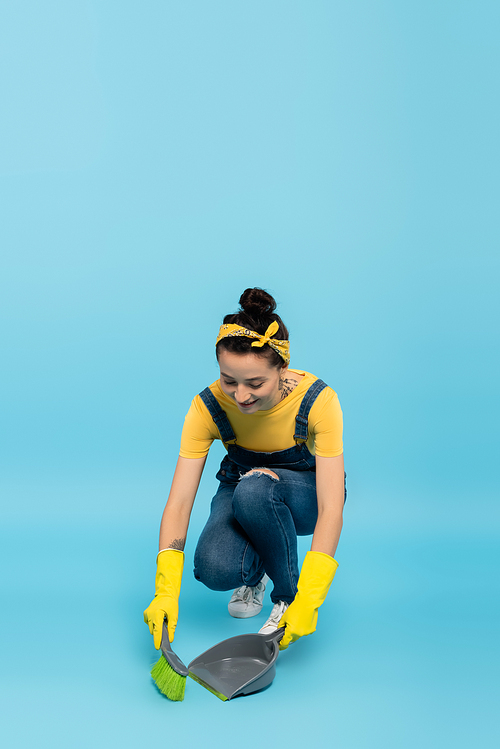 positive housewife in denim overalls and rubber gloves sweeping floor on blue