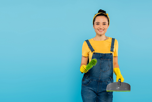joyful housewife with scoop and broom smiling at camera isolated on blue