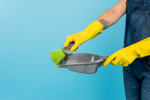 partial view of tattooed housewife in yellow rubber gloves holding scoop and broom isolated on blue