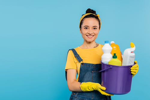 young housewife in yellow rubber gloves holding bucket with detergents isolated on blue