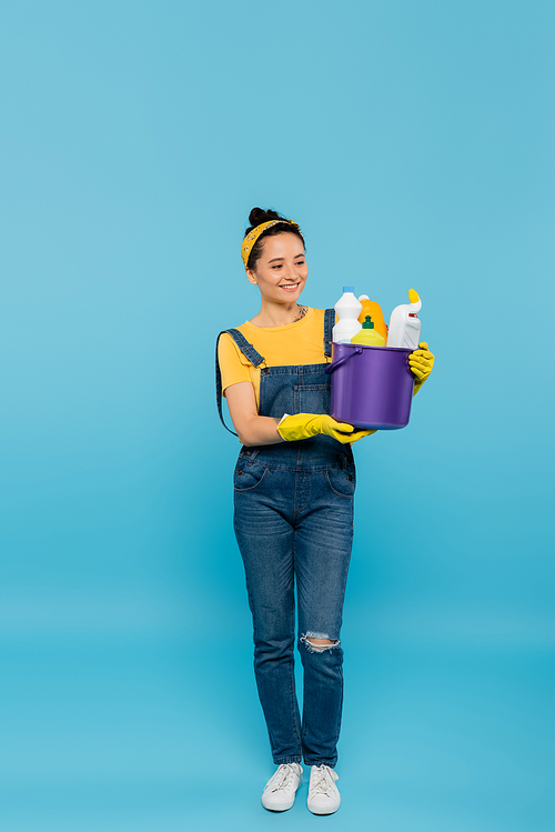full length view of housewife in denim overalls and yellow rubber gloves holding bucket of detergents on blue