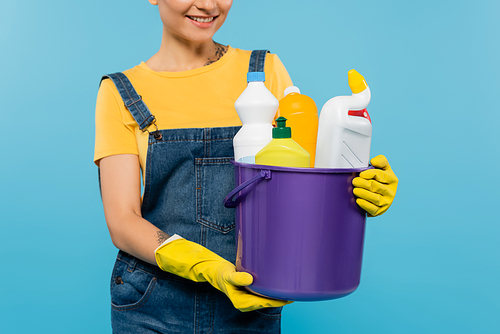 cropped view of smiling housewife in yellow rubber gloves holding bucket with cleansers isolated on blue