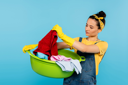 housewife in yellow rubber gloves looking at clothing in laundry bowl isolated on blue