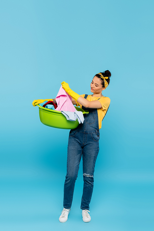 full length view of housewife in denim overalls holding laundry bowl with clothes on blue