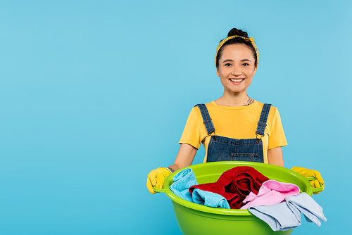 stylish and cheerful woman with laundry bowl smiling at camera isolated on blue
