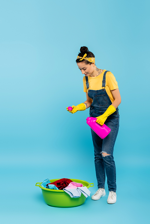young housewife in denim overalls pouring detergent into laundry bowl on blue