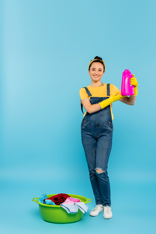 happy housewife in yellow rubber gloves showing bottle of detergent near laundry bowl with clothes on blue