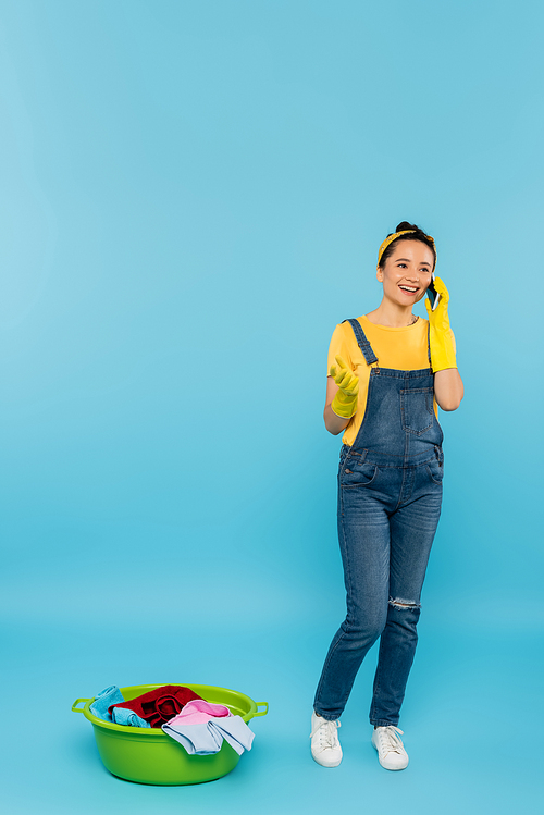 joyful housewife in rubber gloves and denim overalls talking on smartphone near laundry bowl on blue