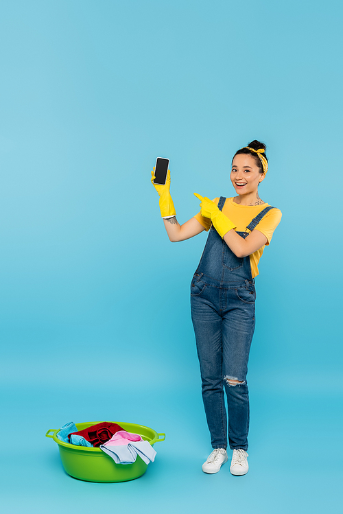 smiling housewife in denim overalls pointing at smartphone near laundry bowl on blue