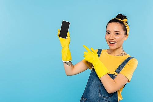 cheerful woman in yellow rubber gloves pointing at smartphone with blank screen isolated on blue