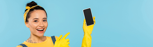 cheerful woman in yellow rubber gloves pointing at laptop with blank screen isolated on blue, banner