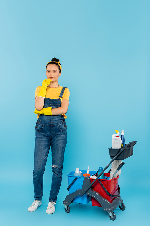 cleaner in rubber gloves and denim overalls  near cart with cleaning supplies on blue
