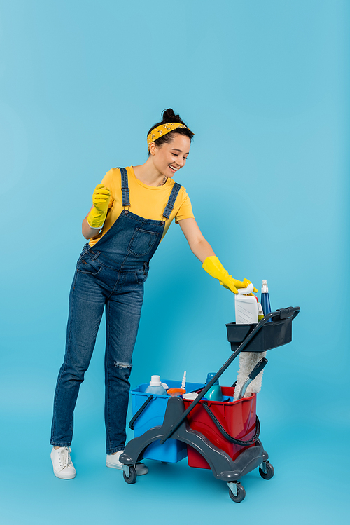 young cleaner in denim overalls smiling near cart with cleaning supplies on blue