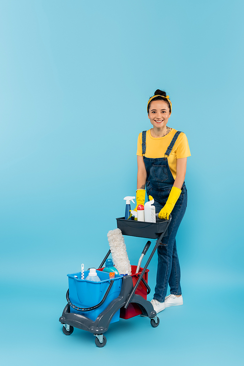 happy cleaner in denim overalls  near trolley with cleaning supplies on blue