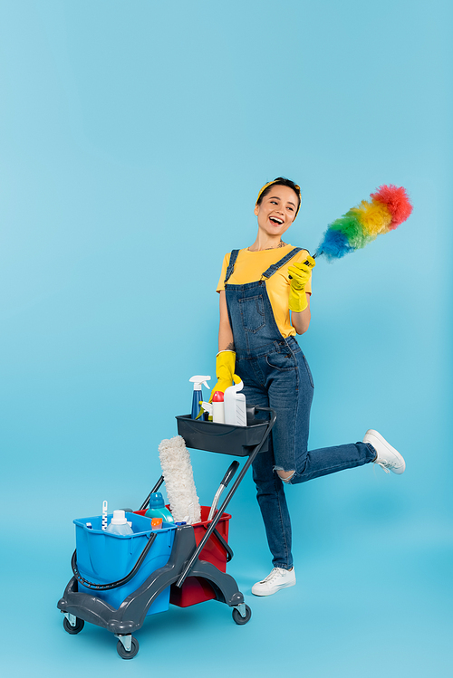 cheerful cleaner with dust brush posing near cart with detergents and buckets on blue