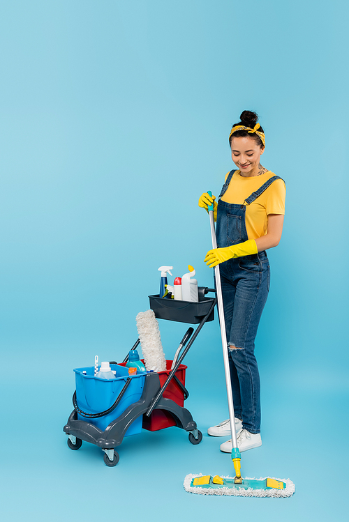 smiling cleaner with mop near cart with buckets and detergents on blue