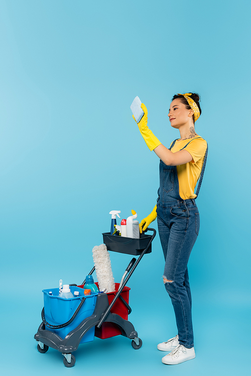 cleaner in denim overalls and rubber gloves taking selfie near cart with cleaning supplies on blue