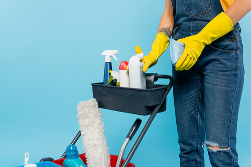cropped view of cleaner in rubber gloves holding sponge near cart with cleaning supplies isolated on blue