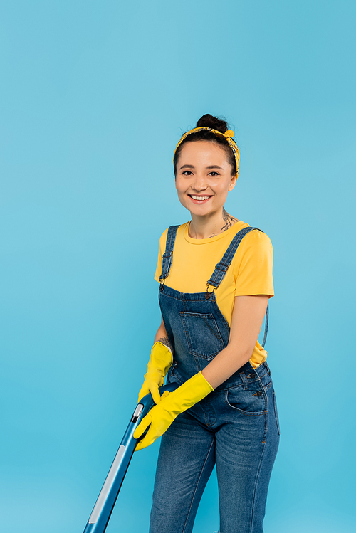 cheerful housewife with vacuum cleaner smiling at camera isolated on blue