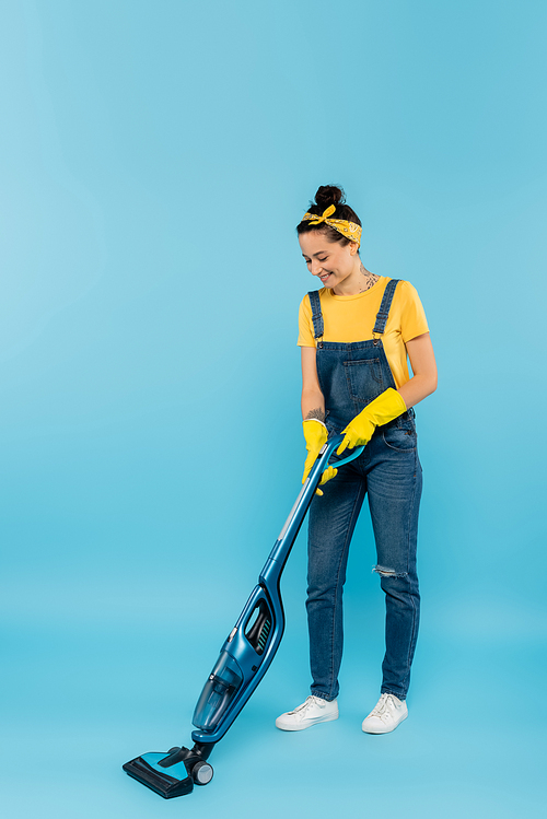 smiling housewife in denim overalls and yellow rubber gloves vacuuming on blue