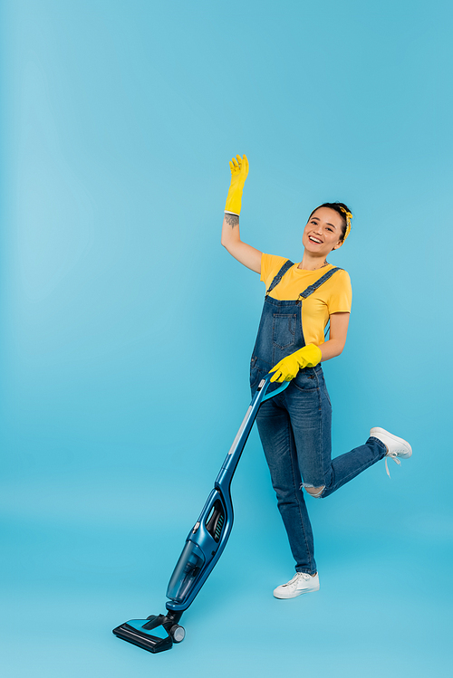 cheerful woman in denim clothes and rubber gloves waving hand while vacuuming on blue