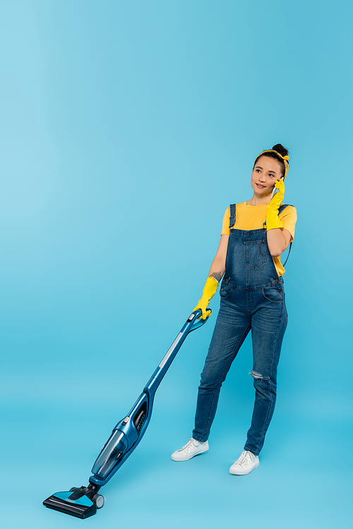 woman in denim overalls and rubber gloves talking on smartphone while vacuuming on blue