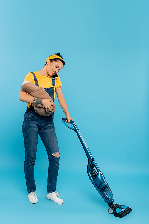 happy woman with baby doll talking on mobile phone while vacuuming on blue