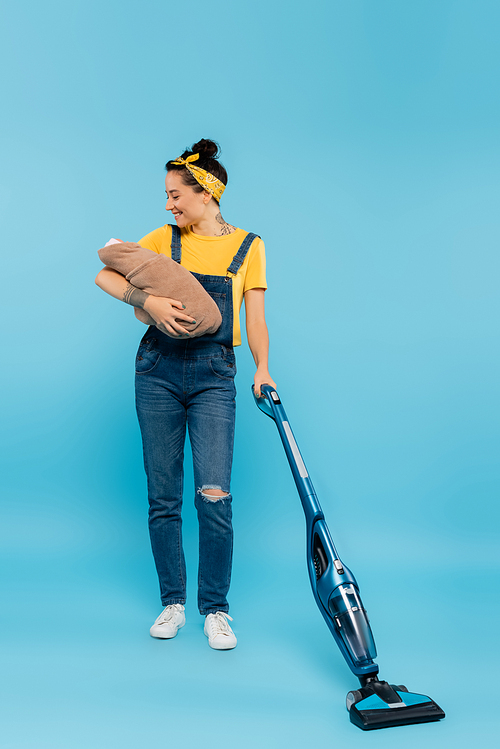 joyful woman in denim overalls holding baby doll while vacuuming on blue